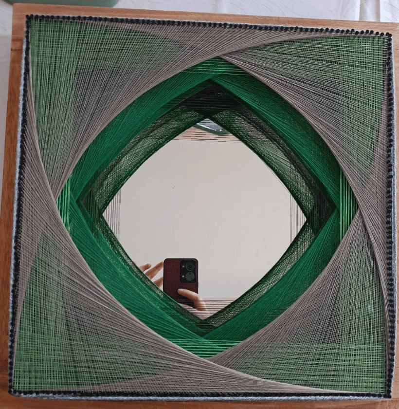 String art piece with green and gray threads interwoven into a symmetrical square pattern, featuring a diamond-shaped hollow center.
