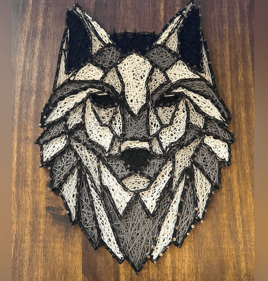 String art wolf head composed of black, white, and gray threads arranged in geometric patterns to create a bold, textured design on a wooden background.